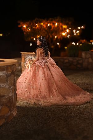 Young woman in a sparkling rose gold quinceañera ball gown holding bouquet, posing outdoors at night with warm string lights.