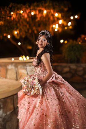Young woman in pink glittery quinceañera gown holding floral bouquet, wearing tiara, posing outdoors at night with warm string lights.