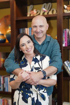 A couple embraces in a warm hug while smiling, with bookshelves visible in the background.