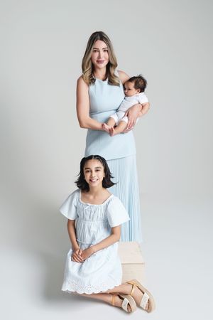 Woman in blue dress holding newborn baby, with young girl in white dress seated on wooden box, studio photo.
