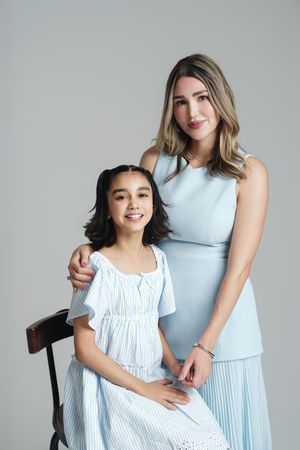 A woman in a light blue dress poses with a smiling young girl in a striped dress seated on a chair.
