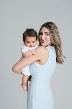 A young woman in a light blue dress holds a baby in a white onesie against a soft gray background.