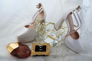 Elegant bridal accessories featuring white platform heels, gold wedding rings, perfume bottles, and delicate white flowers.
