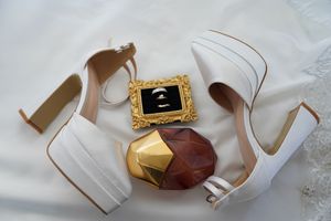 White bridal heels, gold ring frame, and perfume bottle arranged on white fabric for wedding flat lay.