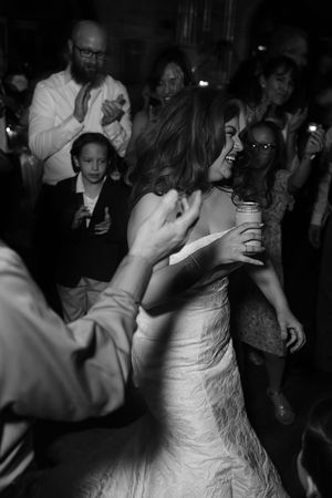 Black and white photo of a bride laughing and dancing with guests at a lively wedding reception.