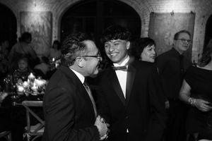 Two men in formal suits smiling and shaking hands at elegant black and white wedding reception.