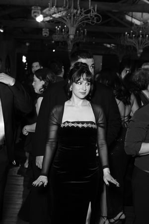 Black and white photo of elegantly dressed woman in velvet gown at formal evening event with chandelier backdrop.