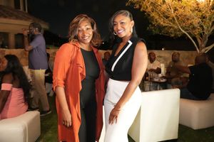 Two women smiling and posing together at an outdoor evening social event with guests mingling in the background.