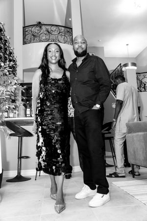 A man and woman pose together in black outfits at an indoor party, captured in black and white photography.