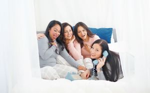 Four women lying together on a white couch share joyful laughter during a casual hangout.