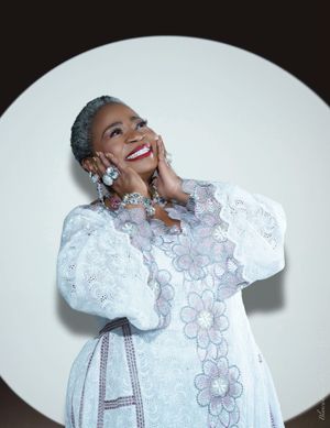 Elegant figure in flowing white dress adorned with floral patterns and puff sleeves poses joyfully against round backdrop.