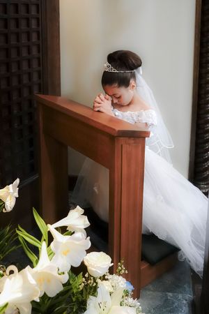 A bride in white lace gown bows in prayer at a wooden altar beside white roses and lilies.