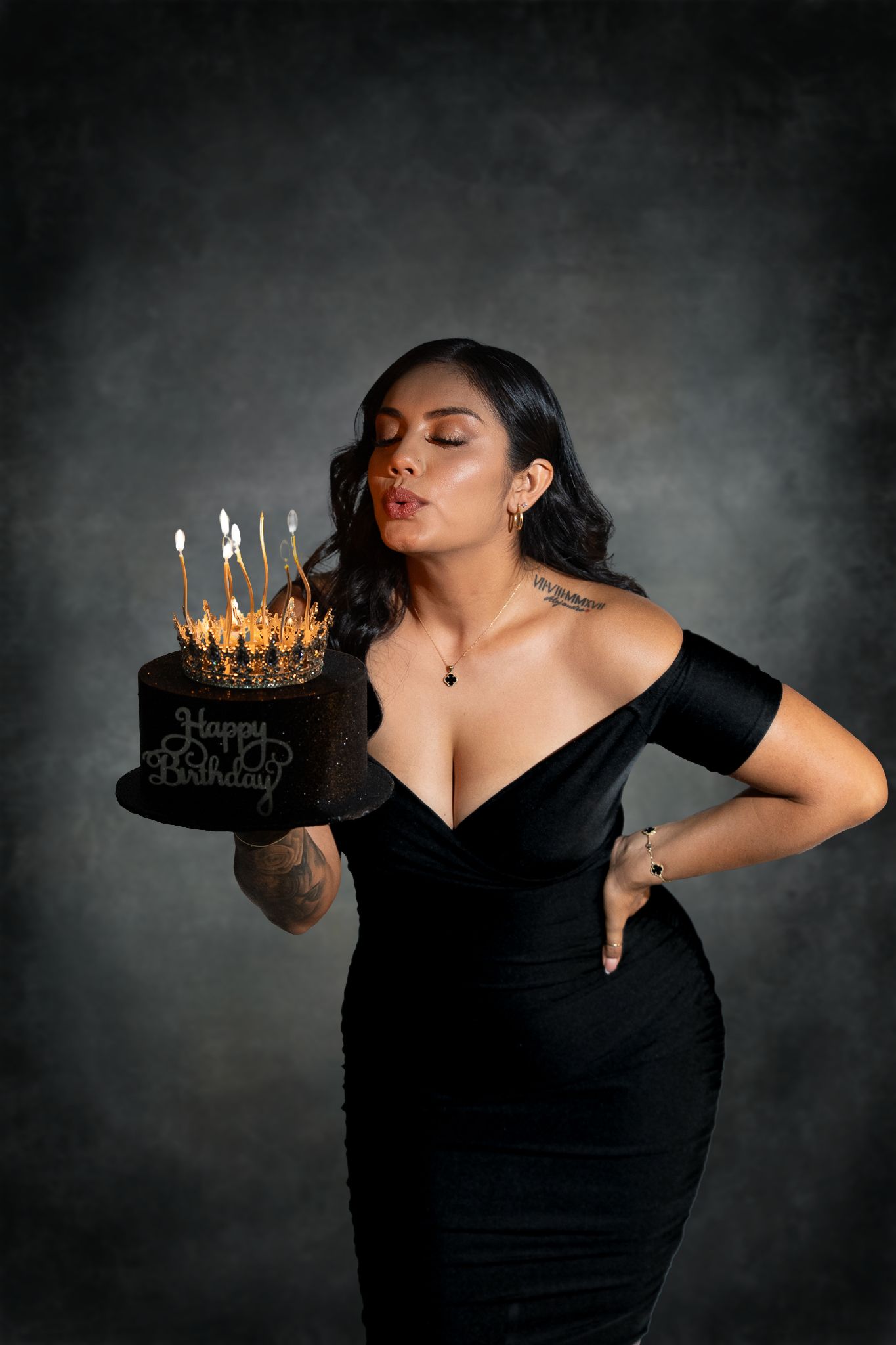 A figure in an elegant black velvet dress blows out candles on a birthday cake against a dark dramatic backdrop.
