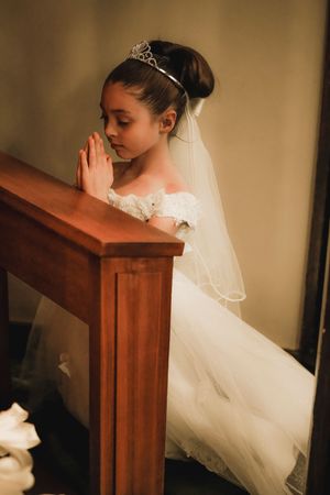 A bride in a white lace dress and veil bows her head in prayer at a wooden church pew.