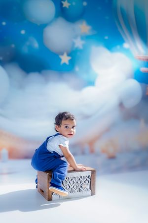 A child in blue sits on a wooden box against a dreamy sky backdrop with bokeh lights and stars.