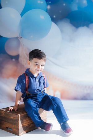 A child in blue overalls sits on a wooden crate surrounded by white and blue balloons in a dreamy studio setting.