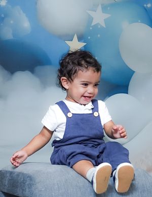 Adorable toddler in navy overalls sitting on a gray cushion against dreamy cloud backdrop with white stars.