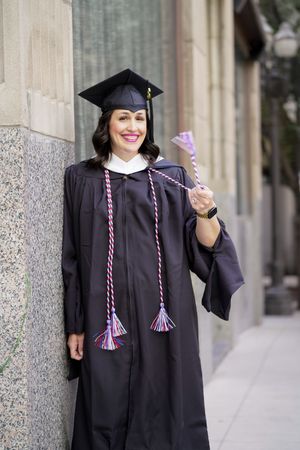 A graduate in traditional black cap and gown holds up their diploma while standing against a stone building exterior.