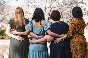 Group of friends wearing colorful dresses stand arm in arm in a row for a casual outdoor photo.