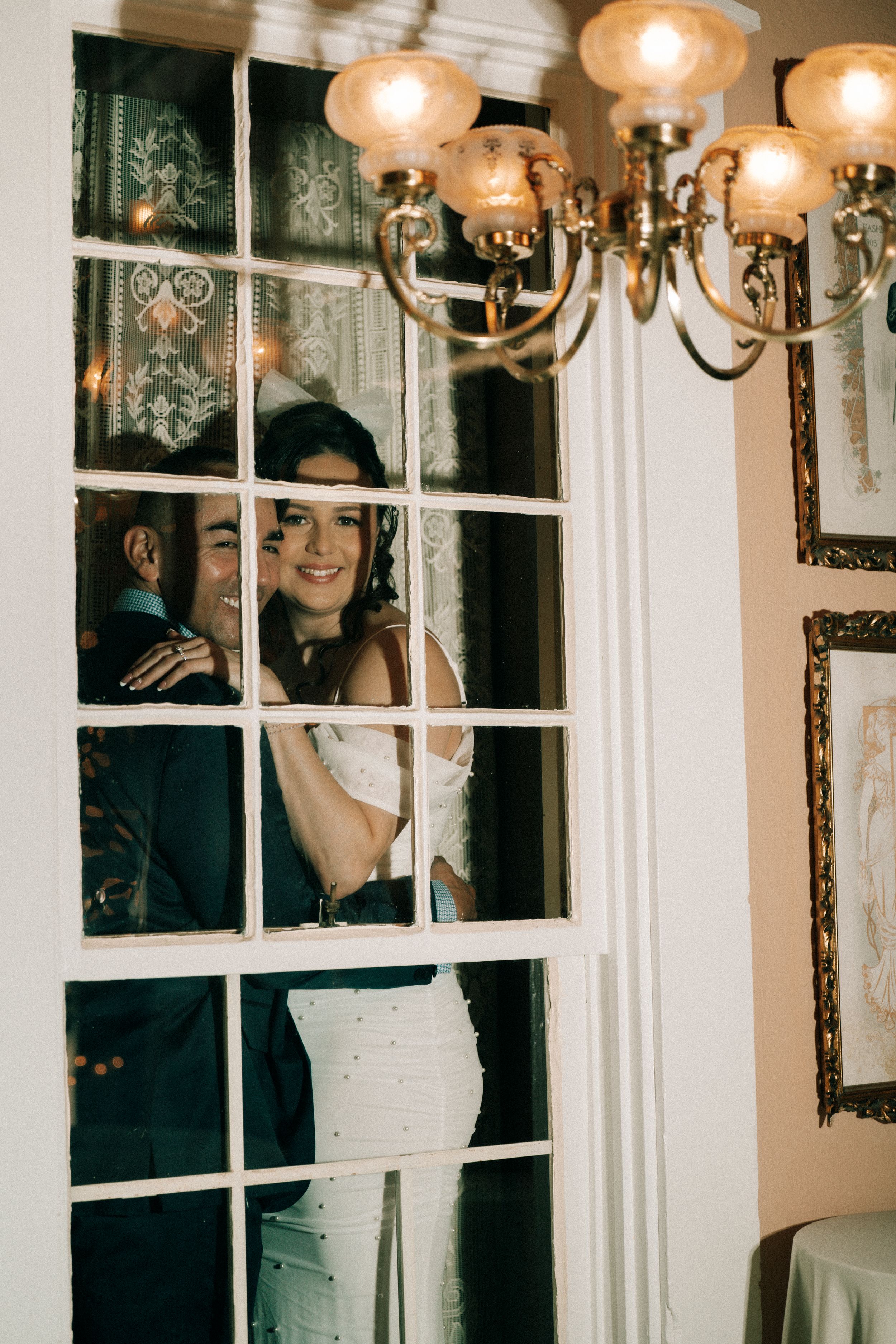 A couple shares a joyful celebration moment under elegant chandelier lighting in a white-decorated venue.