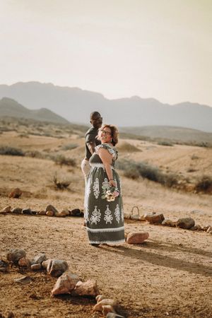Two people embrace while sitting on desert rocks against a backdrop of rolling hills and mountains in warm light.