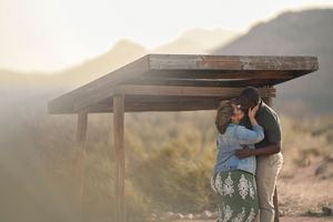 A couple embraces under a wooden shelter at sunset in a desert mountain landscape.