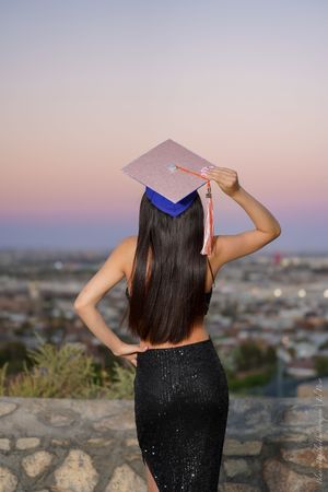 Graduate in black dress and cap poses looking over cityscape during sunset from elevated viewpoint.