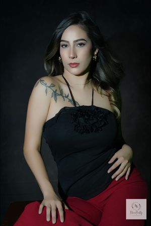 Young woman in black halter top with floral detail and shoulder tattoo poses confidently against dark background.