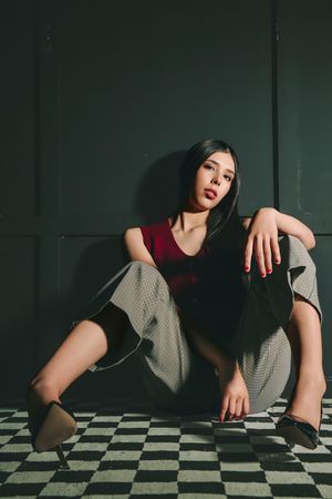 Young Asian woman in red top and plaid pants poses on checkered floor against dark green wall.