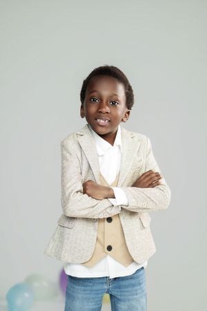 Confident young Black boy in beige blazer and jeans stands with arms crossed, smiling against gray background.