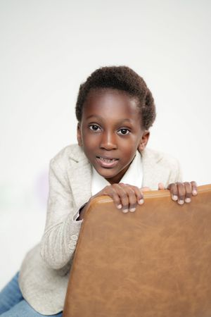 Young Black boy in gray blazer leaning on brown leather chair, smiling at camera against white background.