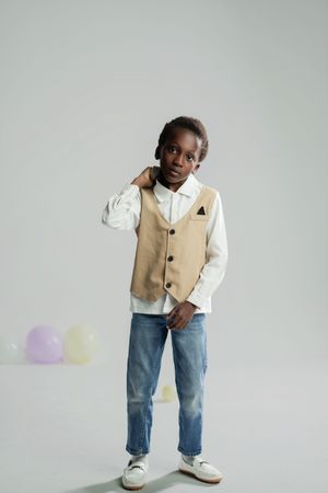 A young Black boy poses confidently in a beige vest, white shirt, and jeans with white sneakers against a gray background.