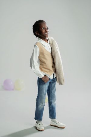 Young boy in white shirt, beige vest, jeans, and loafers poses confidently with hands in pockets on gray background.