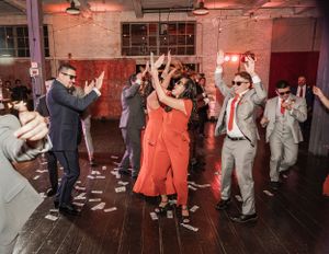 Wedding guests dance enthusiastically on a wooden floor in an industrial venue with exposed brick walls and red lighting.