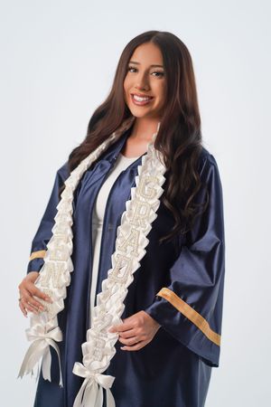 Smiling young woman in navy blue graduation gown with white embroidered sash posing for a graduation portrait.