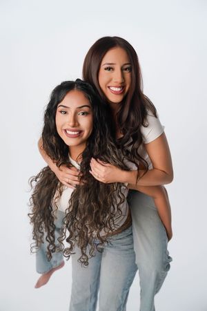 Two smiling young women pose together, one giving the other a piggyback ride against a white background.