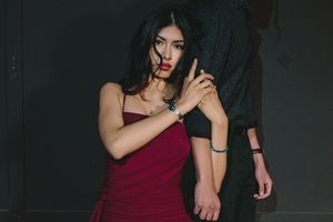 Woman in red dress with red lips holds man's arm in dramatic dark setting.
