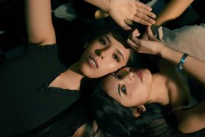 Two young women with dark hair lying down together, faces close, in a dramatic dark-toned artistic portrait.
