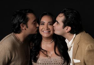 Two men kissing a smiling woman on the cheeks in a warm, affectionate portrait against a dark background.