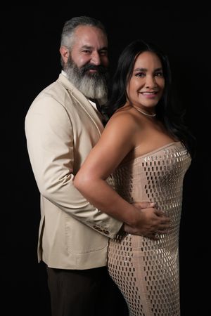 A smiling couple poses together, man in beige blazer with gray beard embracing woman in gold sequin strapless dress.