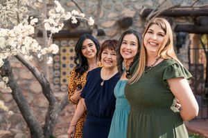 Four friends in colorful dresses smile together in front of blooming cherry blossoms and stone building facade.