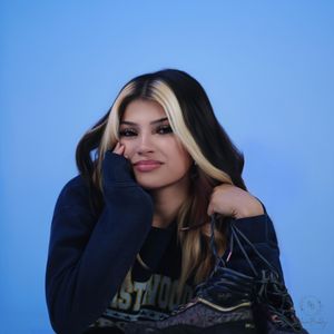 Young woman with black and blonde highlighted hair poses against a blue background wearing a dark graphic sweatshirt.