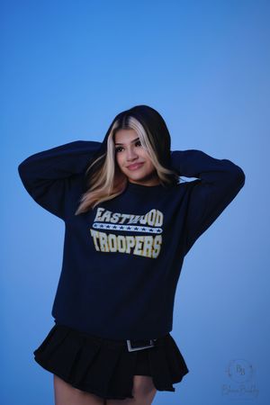 Young woman smiling in Eastwood Troopers navy sweatshirt, posing against blue background.