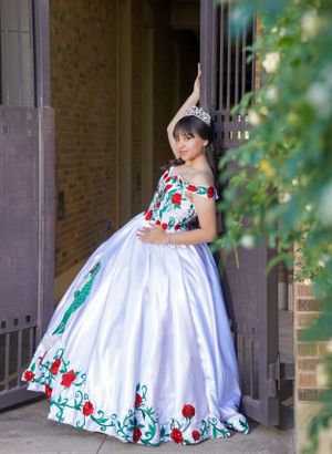 Person in white ball gown with floral embroidery poses gracefully against brick wall.