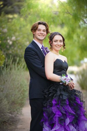 A couple in formal attire poses outdoors, wearing black and purple with a dramatic two-toned wedding dress.