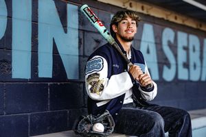Young baseball player in varsity jacket holding bat and glove, posing in dugout for sports portrait photo.