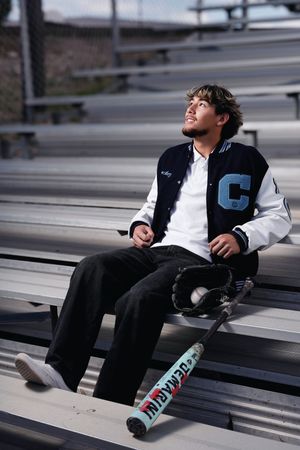 Young man in varsity jacket sits on bleachers with baseball bat and glove, looking up thoughtfully.
