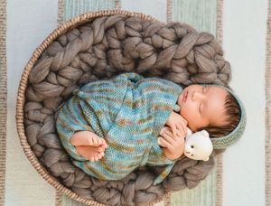 Sleeping newborn baby in a wicker basket with chunky knit blanket, wearing blue striped outfit, holding a small white stuffed animal.