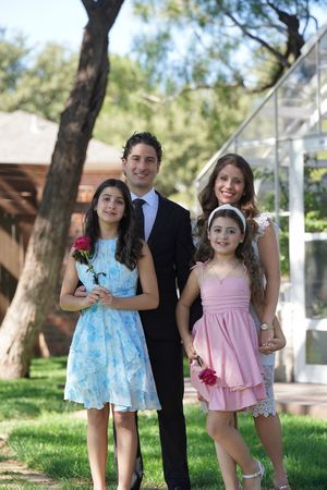 Family of four posing outdoors, two girls in blue and pink dresses holding roses, man in suit, woman smiling.