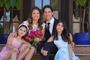 Family of four posing on steps, parents holding pink peonies bouquet, two daughters in formal dresses, outdoor celebration.